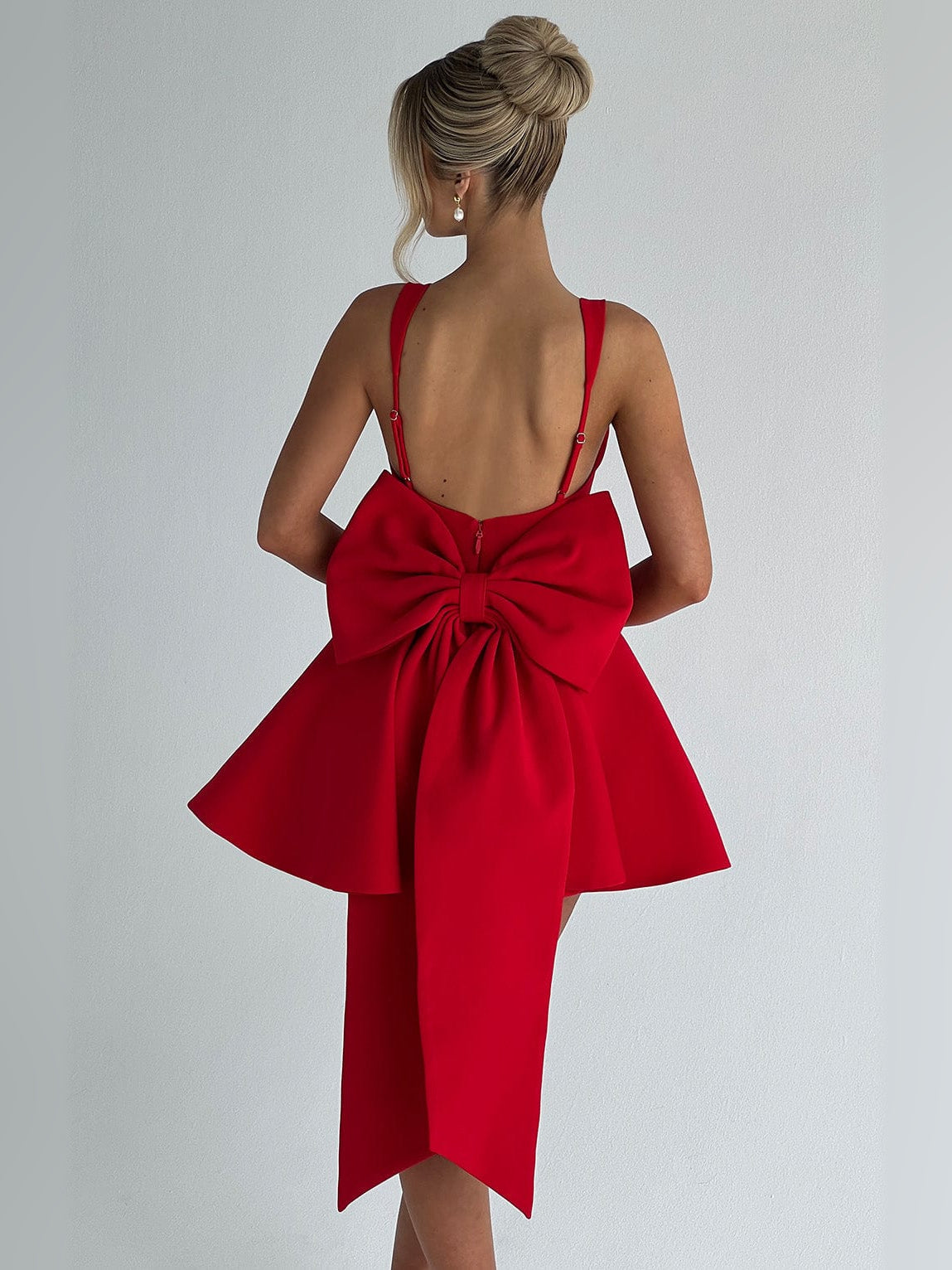 Back view of red sleeveless mini dress with square back, bow at waist, draped tails