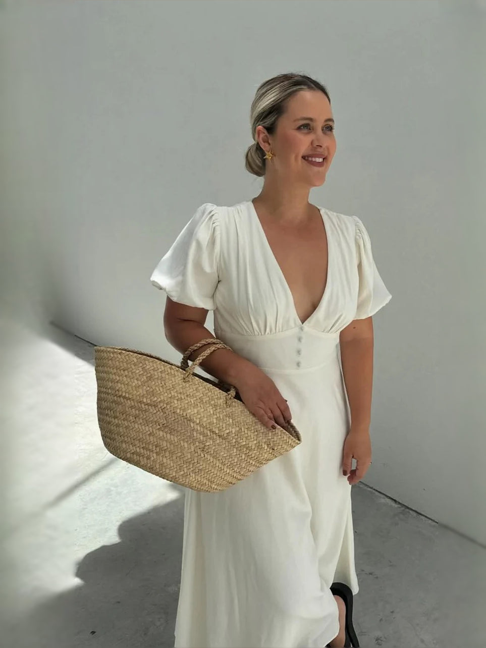 White midi V-neck dress with puff sleeves, held by a model with woven handbag
