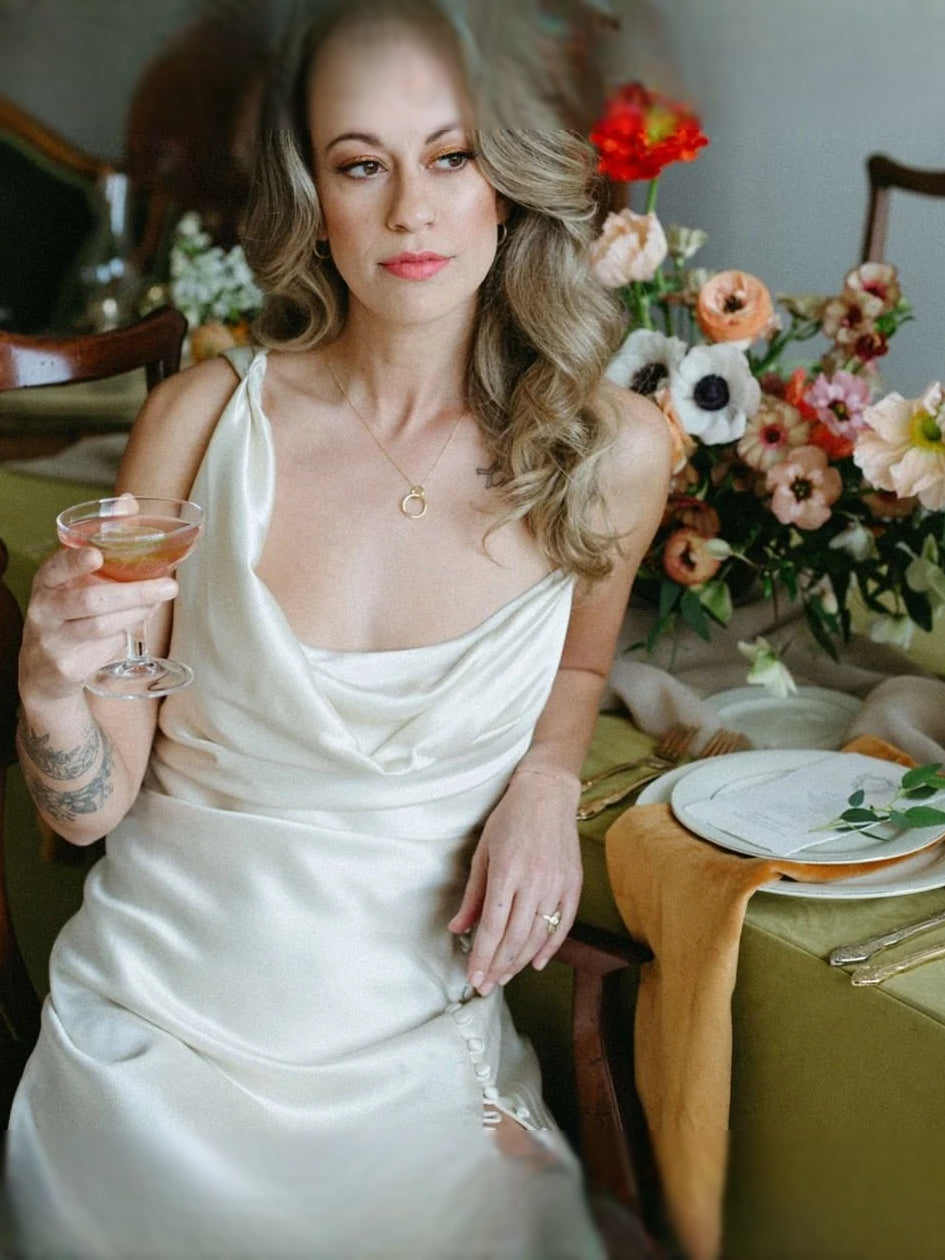 Ivory satin cowl-neck maxi dress with draped corset and thin straps, holding a drink