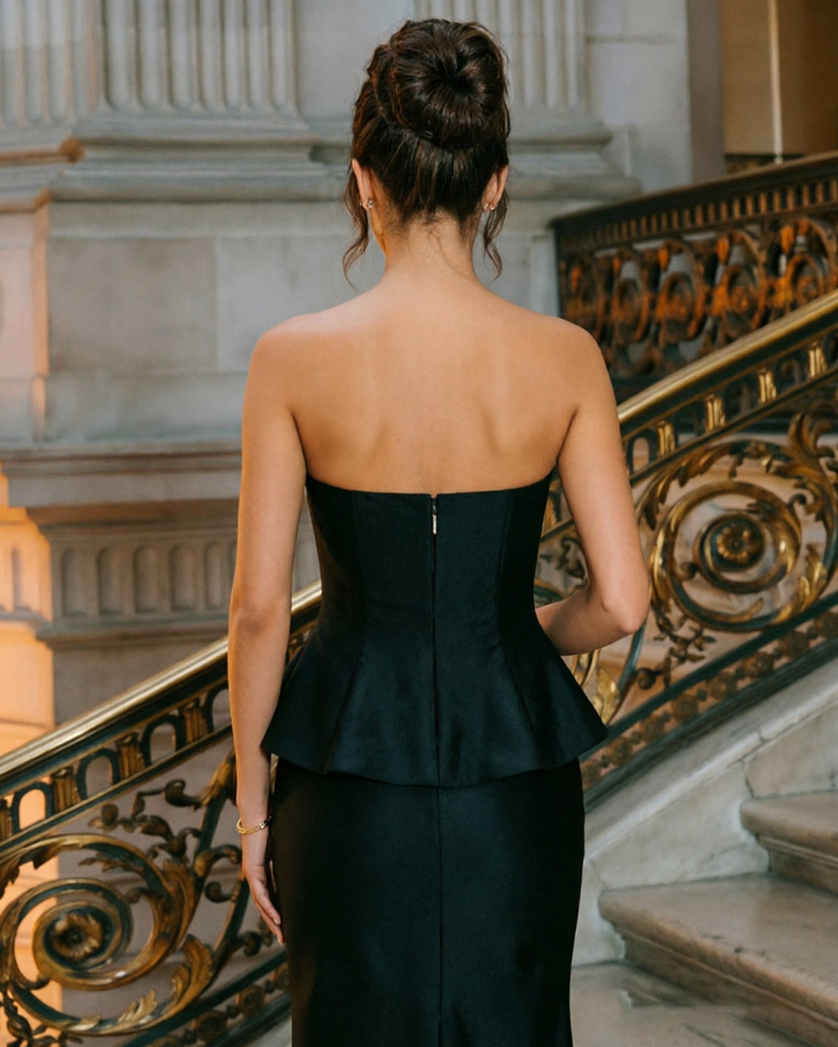 Back view of black strapless peplum mermaid formal dress with satin finish and long skirt.